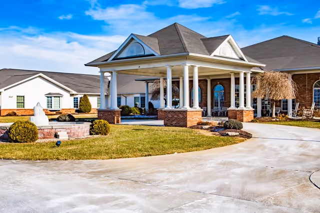 Exterior view of a senior living facility with a covered entrance supported by white columns and brick bases. The building has a combination of brick and white siding with multiple windows. There is a circular driveway and a small landscaped area with bushes and a fountain in front of the entrance under a blue sky with some clouds.