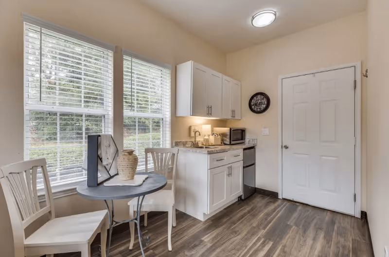 Small kitchenette area with white cabinets, a countertop with a microwave and mini fridge, a round table with two white chairs, and two large windows with blinds letting in natural light. A wall clock is mounted near a closed white door.