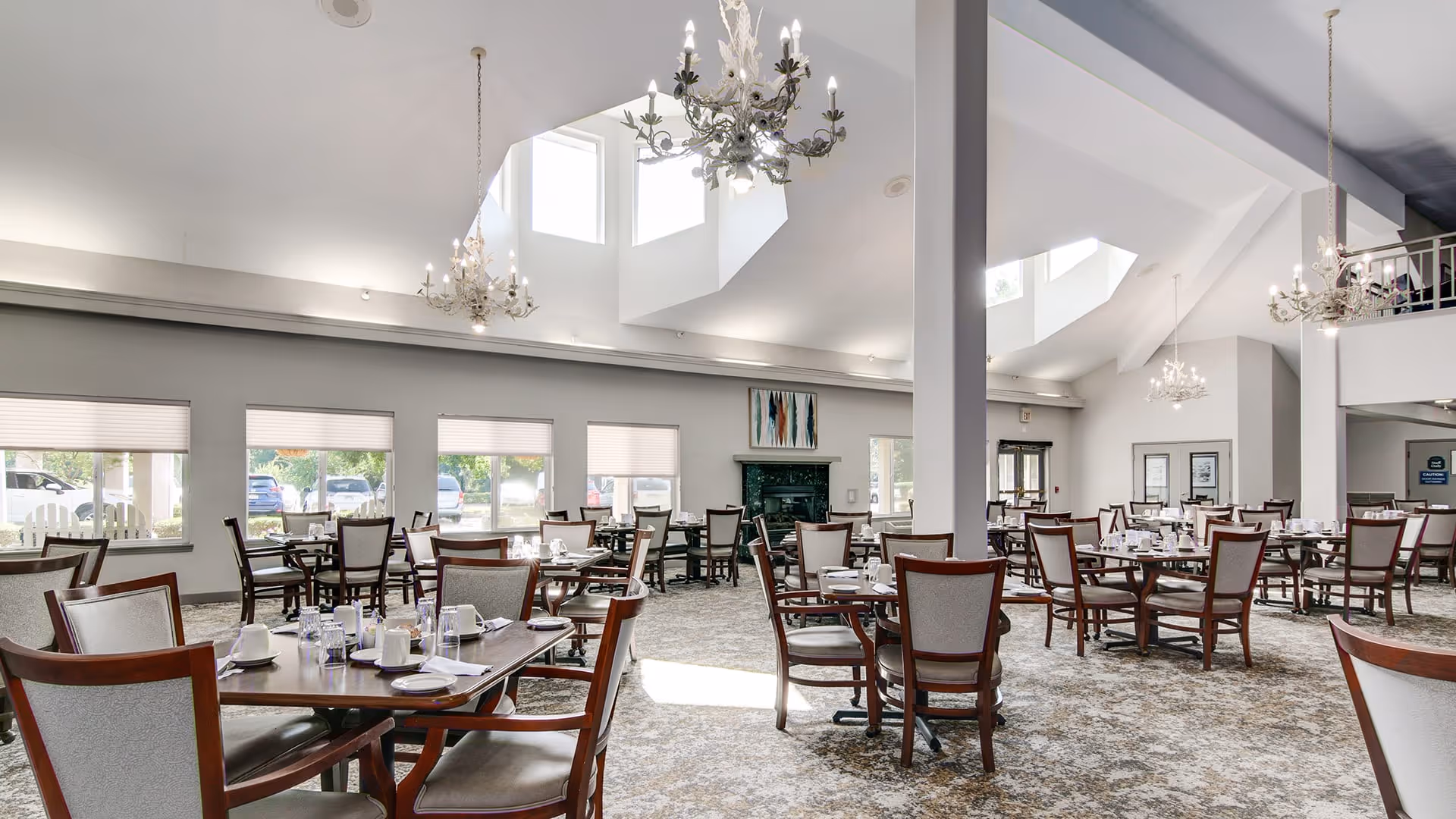 A spacious dining room with multiple wooden tables and chairs arranged neatly. Each table is set with white cups, plates, and glassware. The room features high ceilings with large windows allowing natural light to fill the space. Elegant chandeliers hang from the ceiling, and there is a fireplace along one wall with artwork above it.