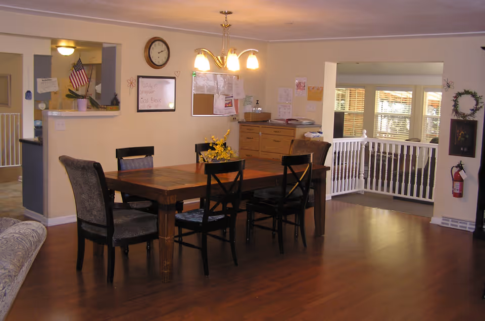 Communal dining area with a wooden table and chairs, bulletin boards on the wall, and a railing opening to a sunlit sitting area.