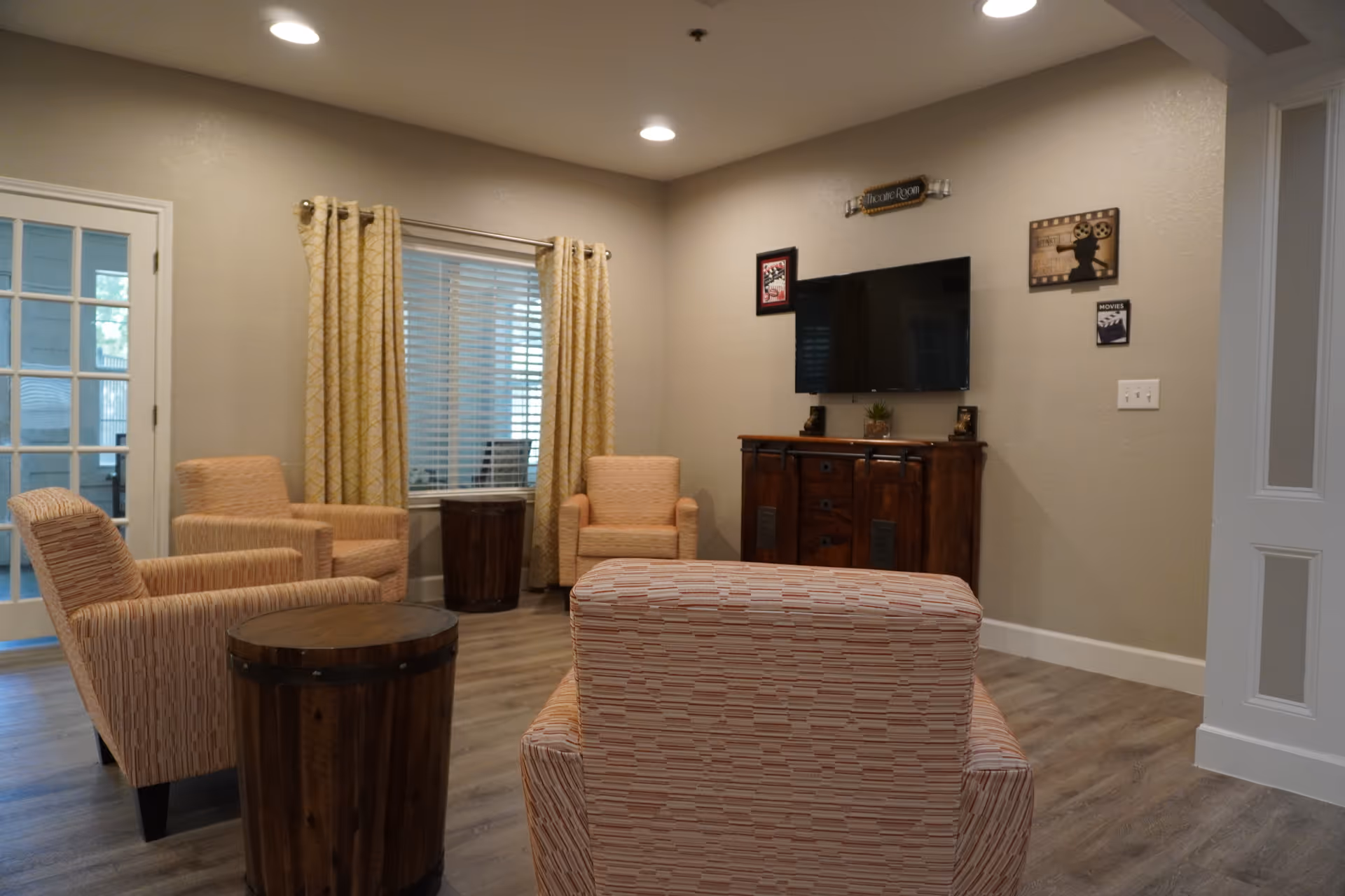 A cozy living room with four patterned armchairs arranged around two round wooden side tables. The room has wood flooring, beige walls, and a window with yellow patterned curtains. A flat-screen TV is mounted on the wall above a wooden cabinet, and there are decorative movie-themed wall hangings nearby. A glass door is visible on the left side of the room.
