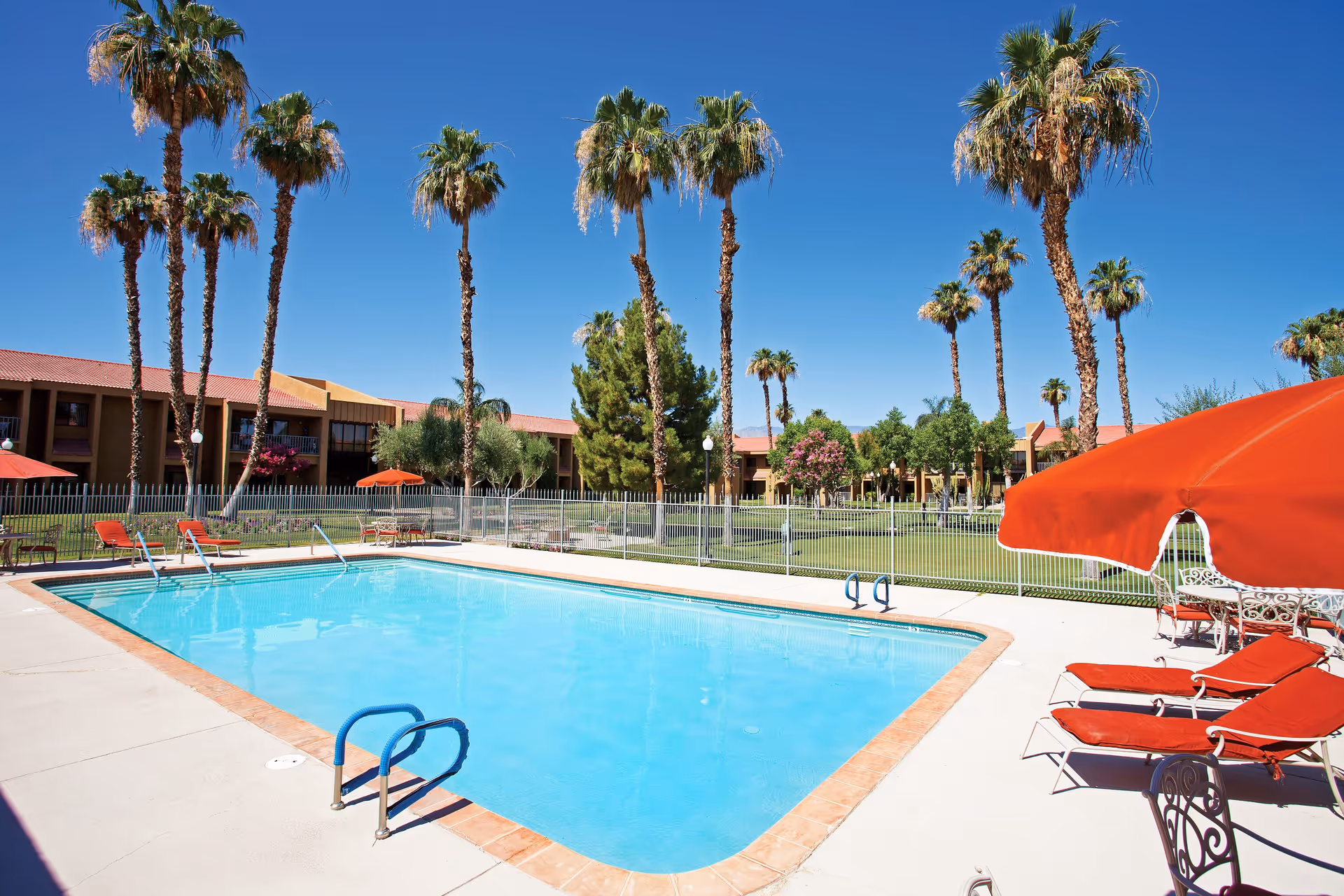 Outdoor swimming pool with clear blue water surrounded by a concrete deck. Several red lounge chairs and umbrellas are arranged around the pool. Tall palm trees and other greenery are visible in the background along with a two-story building under a clear blue sky.