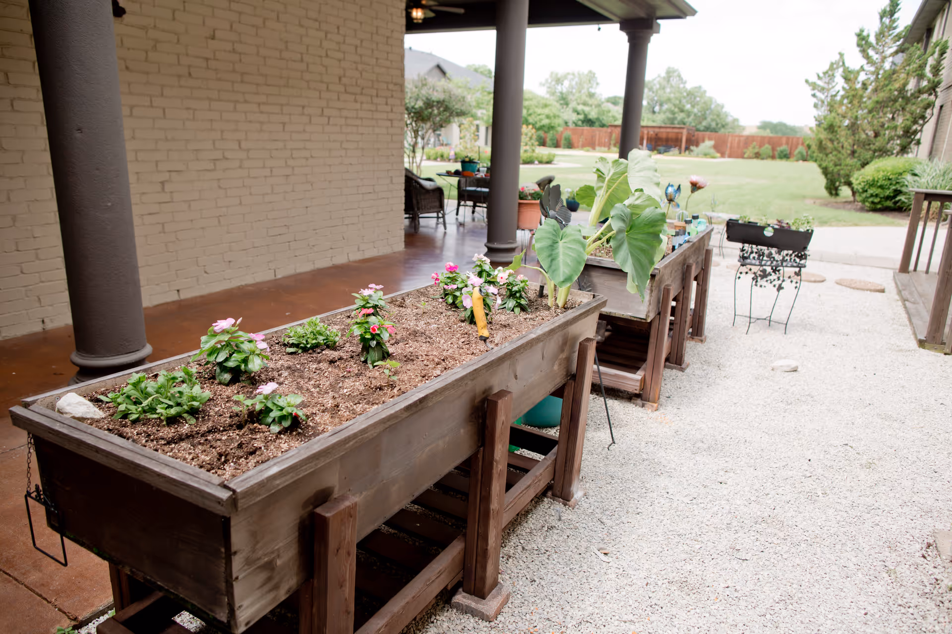 Raised wooden planters with flowering plants on a gravel patio under a covered porch overlooking a grassy yard.