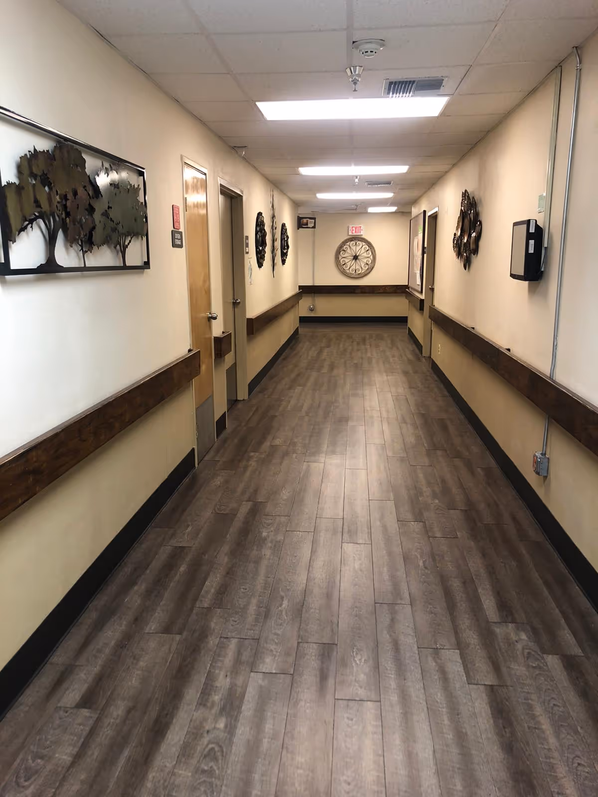 A long, empty hallway inside a healthcare facility with wood-look flooring, beige walls, and ceiling lights. The walls are decorated with metal art pieces and there are several closed doors along the hallway. A large decorative clock is mounted on the far wall beneath an exit sign.