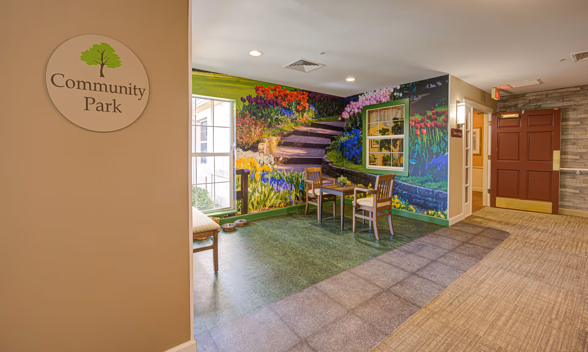 Indoor seating area with two wooden chairs and a small table on green flooring, next to a wall mural depicting a colorful garden with flowers and stone steps. A window with a green frame is part of the mural. A sign on the left wall reads 'Community Park'. The hallway has carpeted flooring and a red door at the end.