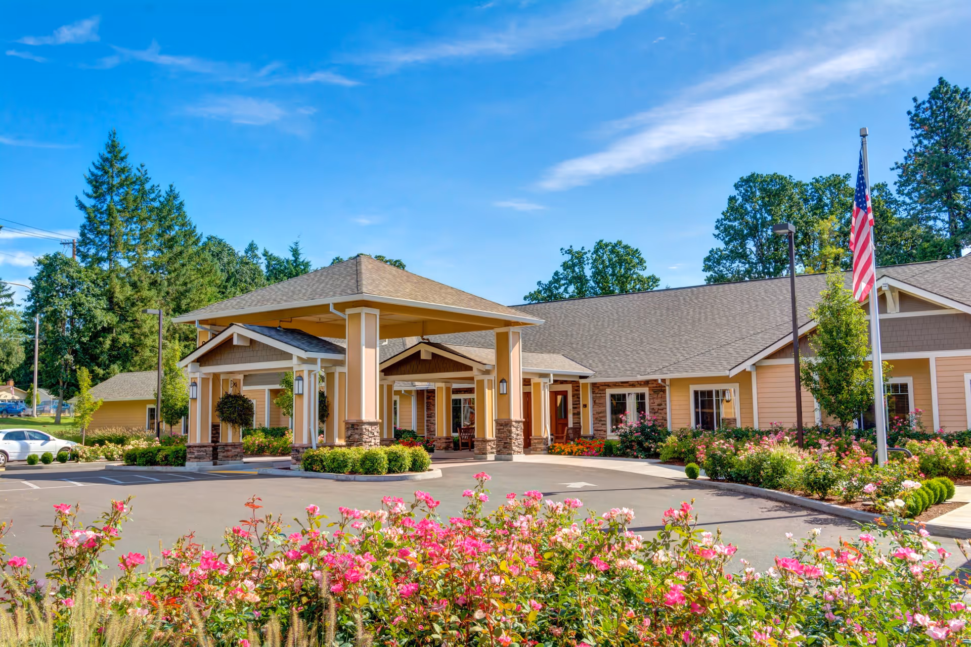 Exterior view of Washington Gardens Memory Care facility on a sunny day, featuring a covered entrance with stone pillars, landscaped flower beds with pink flowers, an American flag on a flagpole, and surrounding trees under a clear blue sky.