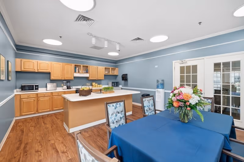 A bright kitchen and dining area with light wood cabinets, white countertops, and blue walls. The kitchen features appliances including a microwave, toaster, and coffee maker. In the foreground, there is a dining table covered with a blue tablecloth and decorated with a colorful floral arrangement. The room has wood flooring and recessed lighting, with double glass doors on one side.