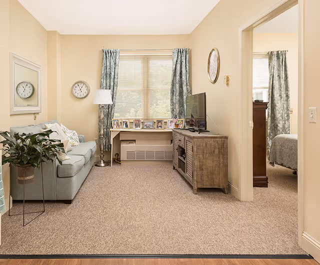 A cozy living room with beige walls and carpeted floor. The room features a light blue sofa with cushions on the left, a tall floor lamp beside it, and a large window with blue patterned curtains in the center. Below the window is a shelf displaying several framed photos. On the right side, there is a wooden TV stand with a flat-screen television. A doorway on the right leads to a bedroom with a bed and matching curtains.