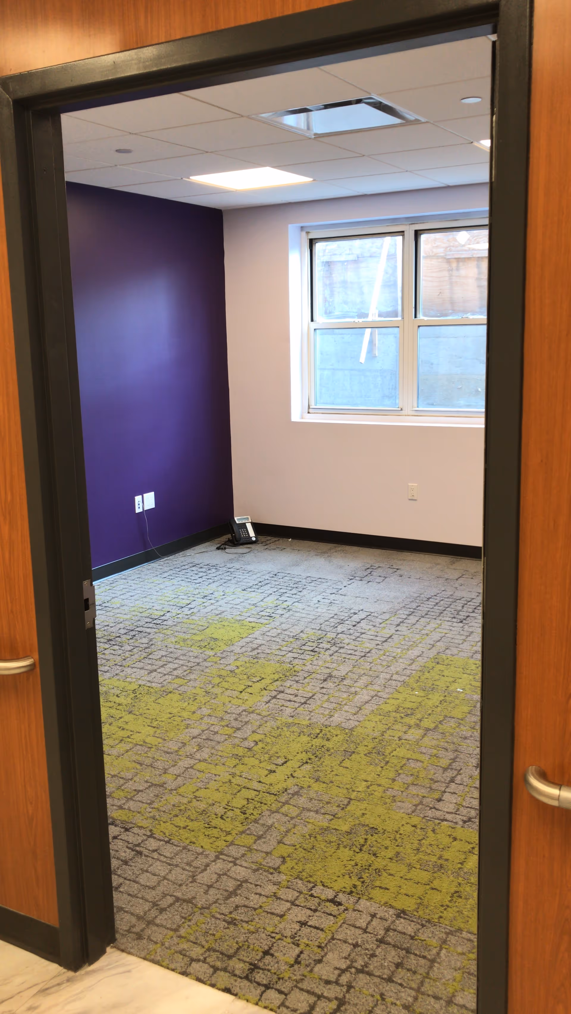 View through a doorway into a small empty room with a purple accent wall and a window. The floor is carpeted with a pattern of gray and green squares. A telephone is placed on the floor near the corner under the window.