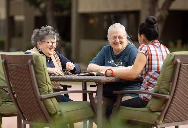 Three women sitting around a table outdoors, engaged in conversation and smiling. The setting appears to be a patio area with cushioned chairs and a building in the background.