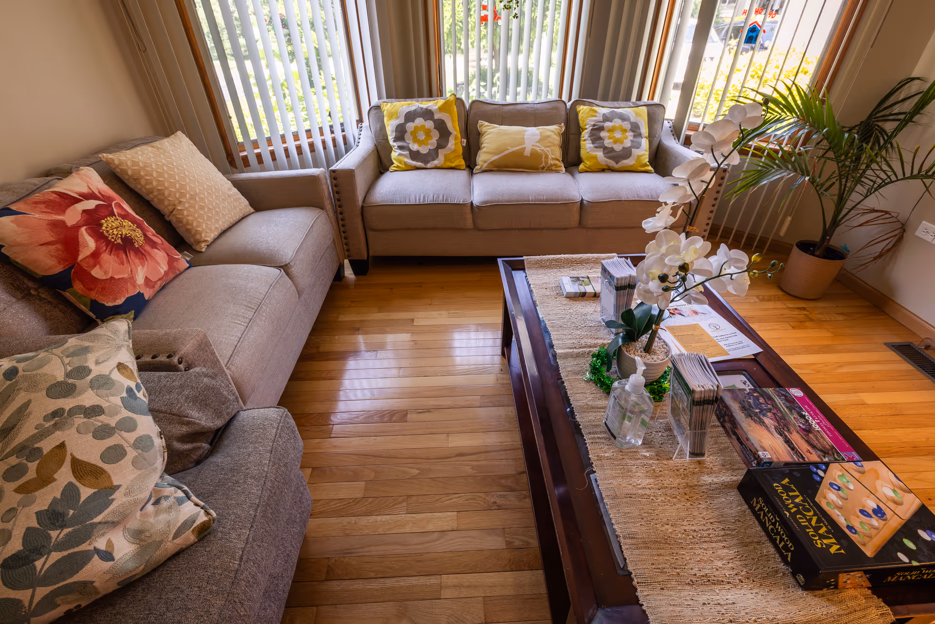 A cozy living room with two beige sofas adorned with colorful patterned cushions. A wooden coffee table in the center holds a potted white orchid, hand sanitizer, brochures, and board games. The room has wooden flooring and large windows with vertical blinds letting in natural light. A potted plant is placed near the window.