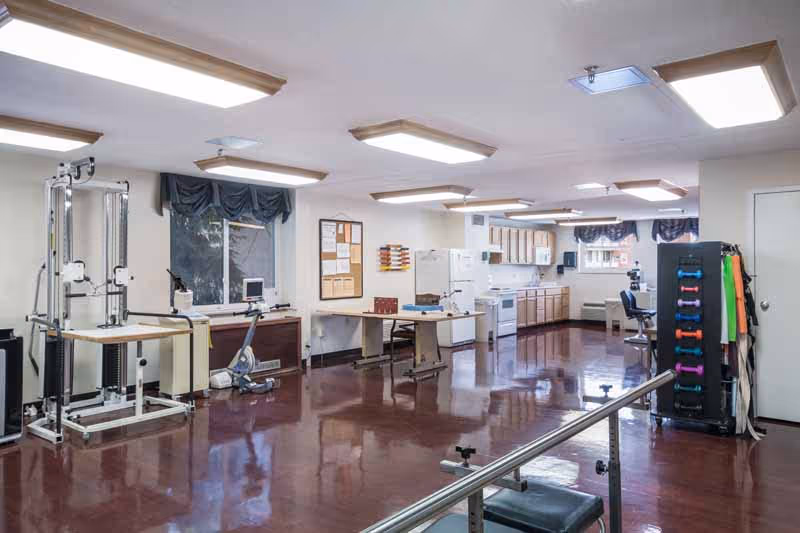 A spacious room with exercise equipment including a stationary bike, weight machine, and a rack of colorful dumbbells. The room has a shiny maroon floor, fluorescent ceiling lights, windows with dark valances, and a kitchenette area with a refrigerator, stove, and cabinets. There are also tables and chairs along the walls.