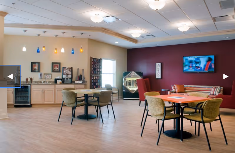 A spacious common area with light wood flooring, featuring two tables with chairs, a small kitchenette with cabinets, a coffee machine, and a mini fridge. The back wall is painted dark red and has a mounted flat-screen TV showing a sports game. There is a striped couch with pillows and two red armchairs near the TV, and a window letting in natural light.