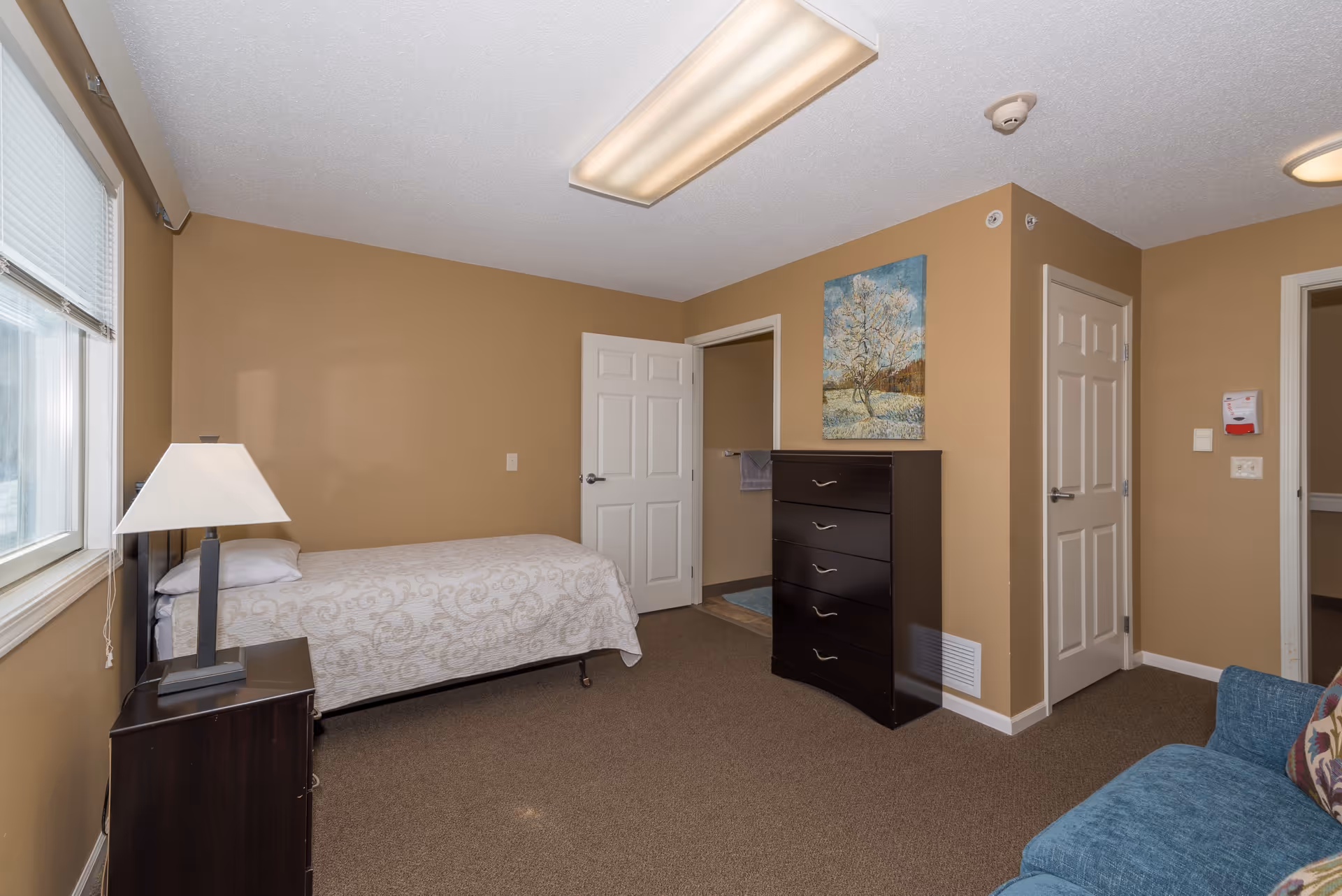 A simple bedroom with beige walls and carpet. The room contains a single bed with a patterned bedspread, a dark wooden nightstand with a lamp, a dark wooden chest of drawers, and a blue upholstered chair with a decorative pillow. There is a window with blinds on the left wall and two white doors on the far wall, one leading to a bathroom. A painting of a tree is hung above the chest of drawers.