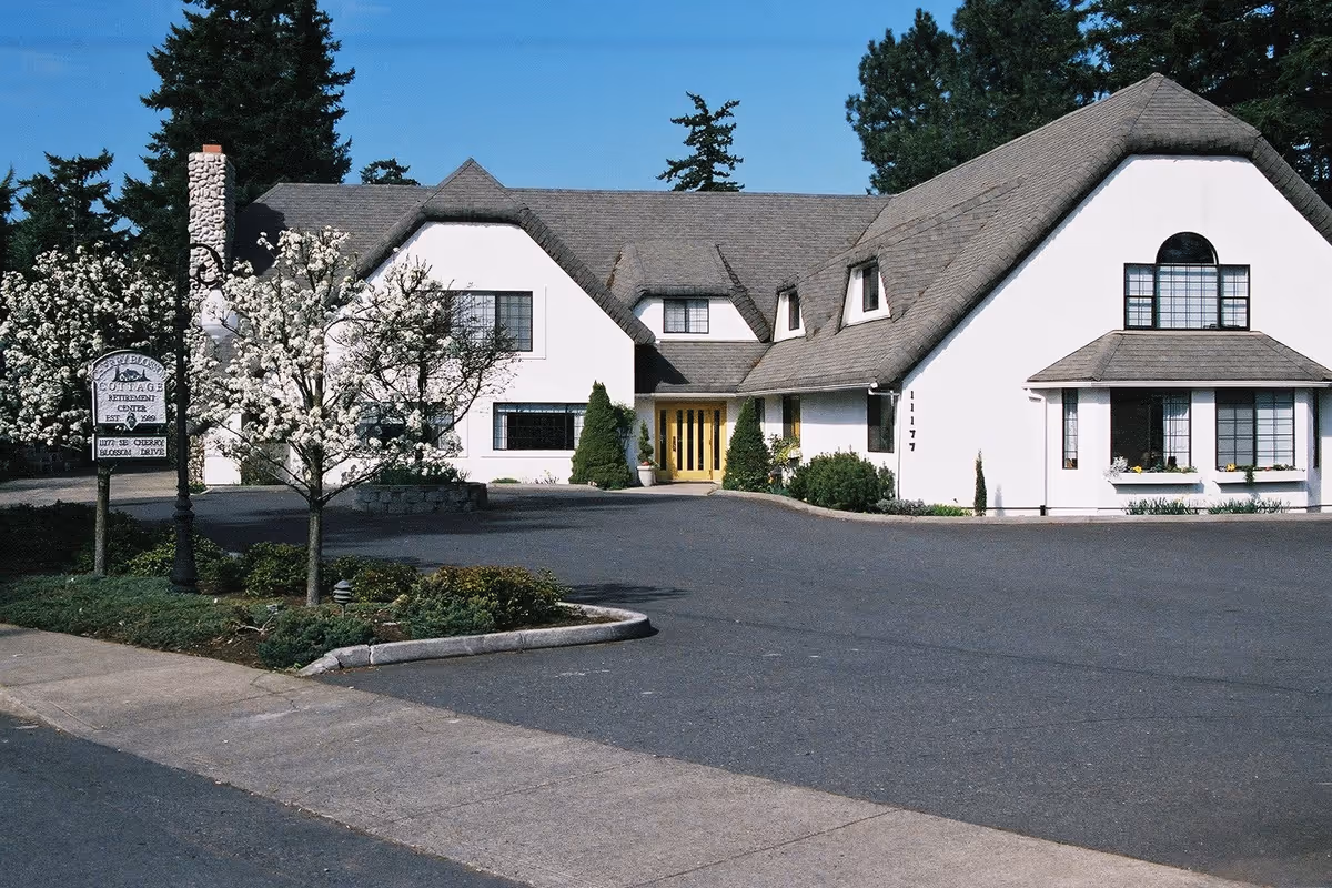 Exterior view of Cherry Blossom Cottage, a residential care facility for seniors, showing a large white building with a gray roof, multiple windows, and a yellow front door. There are blooming trees and shrubs in front of the building, a paved driveway, and a sign with the facility's name near the sidewalk.
