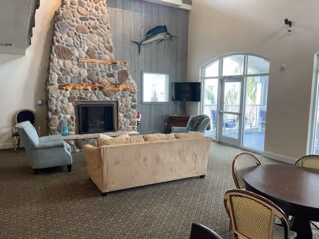 A cozy living room area with a large stone fireplace, a beige sofa, two armchairs, a flat-screen TV mounted on a wooden cabinet, and a round dining table with chairs. There is a large arched window and glass door leading to an outdoor patio with palm trees visible outside. A mounted fish decoration is on the wall above the TV.