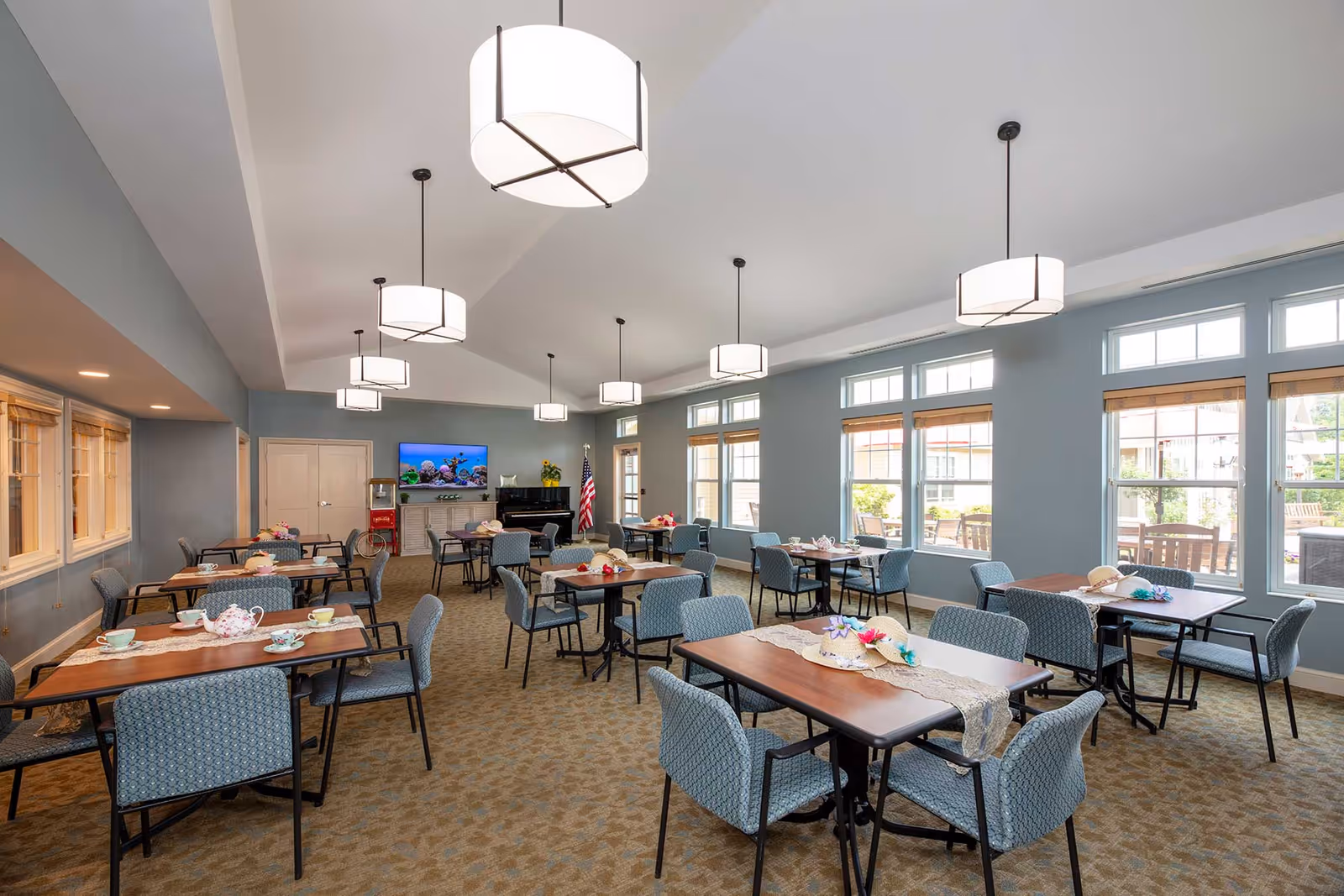 A spacious dining room with multiple wooden tables and blue upholstered chairs arranged neatly. Each table is decorated with a table runner and small floral centerpieces. Large windows on one side allow natural light to fill the room. Modern pendant lights hang from the ceiling. A television and an American flag are visible at the far end of the room.