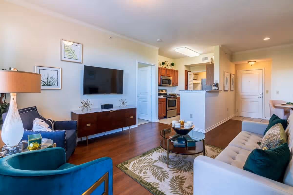 Bright open-plan living room with sofa and chairs facing a wall-mounted TV, a coffee table, and a view into the kitchen and entryway.