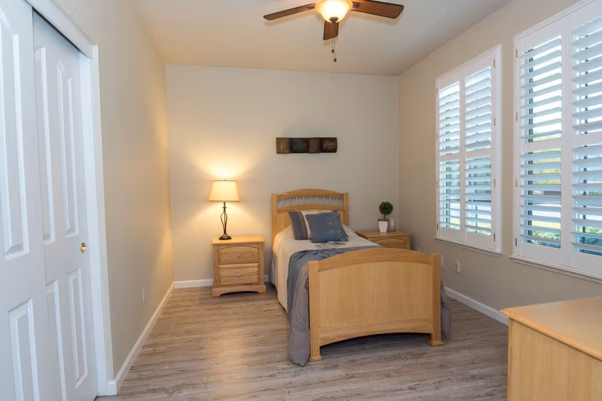 A simple, clean bedroom with a single wooden bed, a nightstand with a lamp, a small plant on another nightstand, and large windows with white shutters letting in natural light.