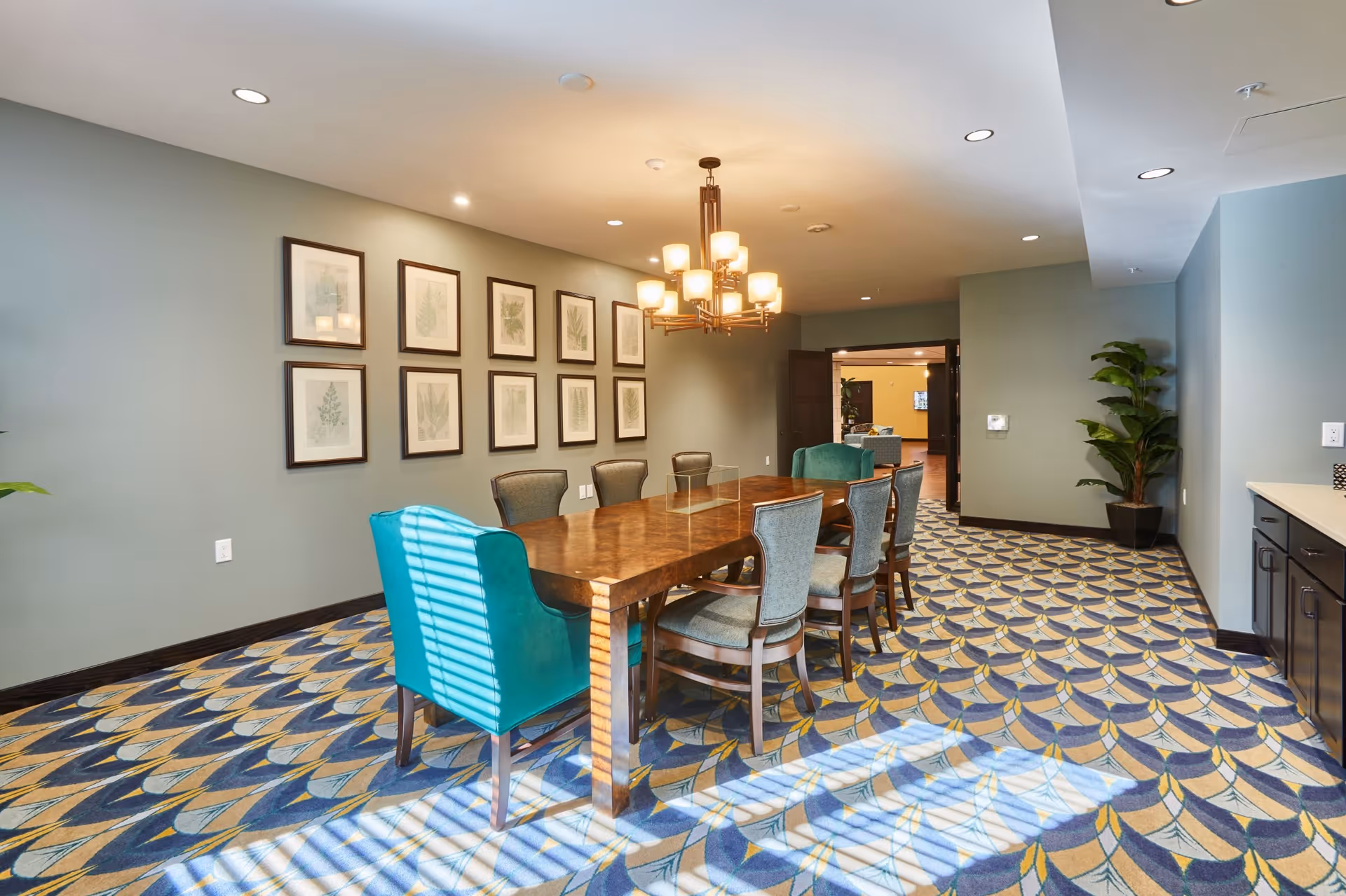 Bright dining/meeting room with a long wooden table surrounded by upholstered chairs, teal accent chairs, framed botanical prints on the wall, and patterned carpet.