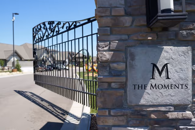 Stone entrance pillar with a plaque reading 'The Moments' beside an open wrought-iron gate at the facility entrance.