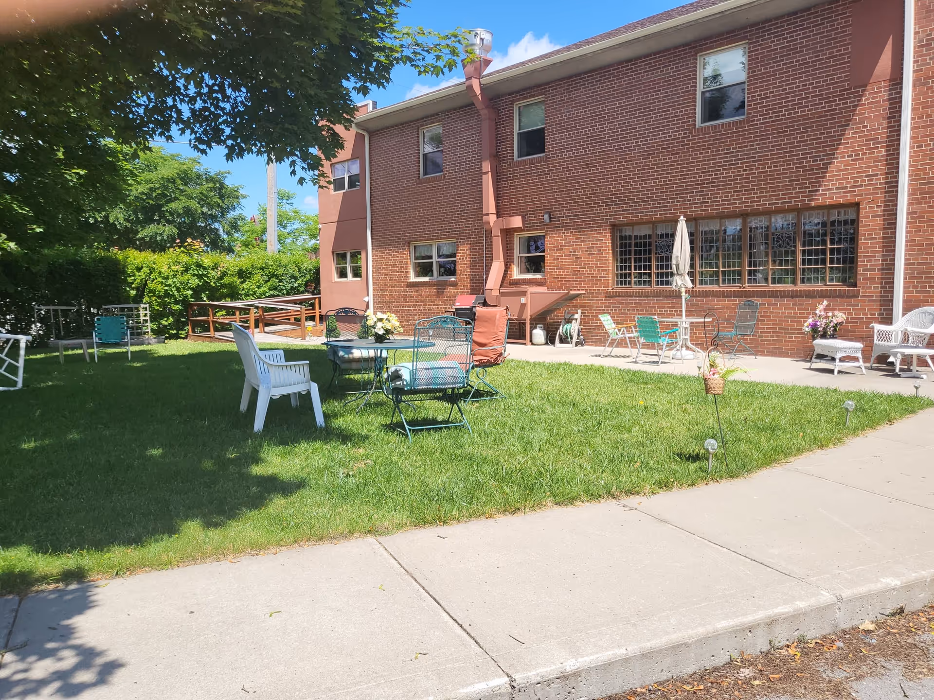 Outdoor patio area of a senior living facility with green grass, several chairs and tables, a brick building in the background, and a tree providing shade.