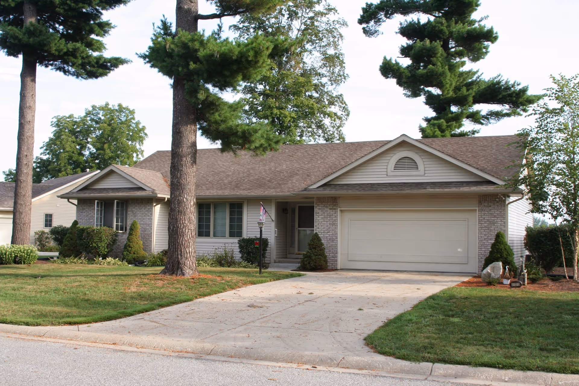 Single-story residential building with a two-car garage, beige siding, and brick accents. The house is surrounded by green grass, trees, and shrubs, with a concrete driveway leading to the garage. The sky is clear with some clouds.