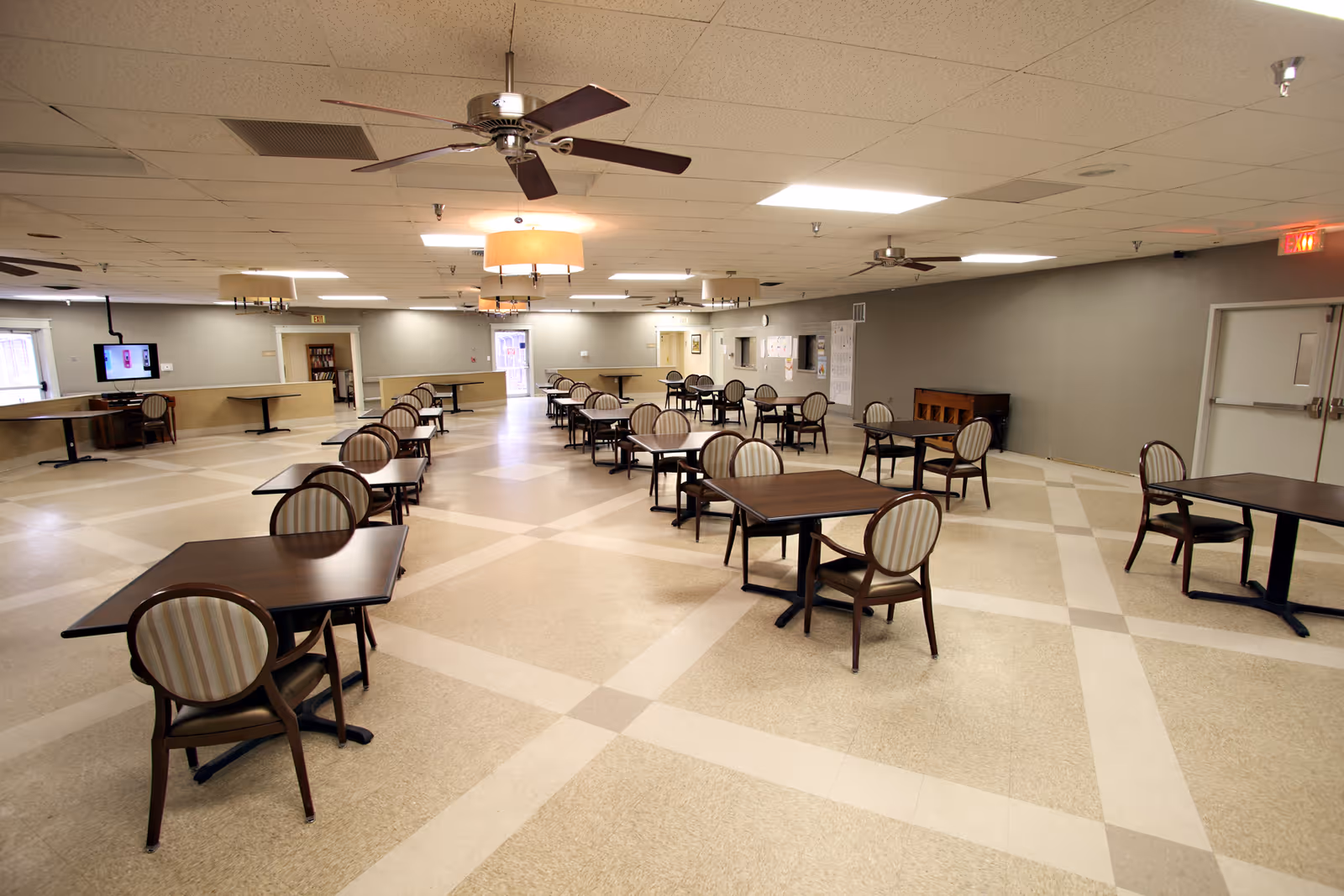 Spacious dining room with multiple square tables and striped upholstered chairs arranged in rows under ceiling fans and pendant lights.