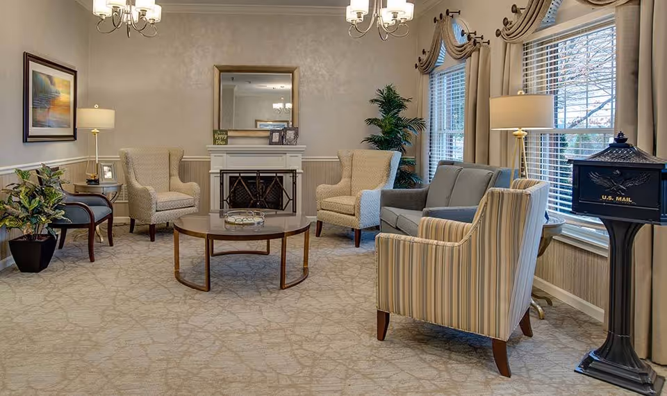 A cozy senior living facility common area with a beige carpet and neutral-colored walls. The room features a round wooden coffee table in the center, surrounded by two beige armchairs, a gray loveseat, a striped armchair, and a wooden chair with blue upholstery. There is a white fireplace with a mirror above it, two floor lamps, a potted plant, and large windows with beige curtains letting in natural light. A black U.S. Mail mailbox stands near the window on the right.