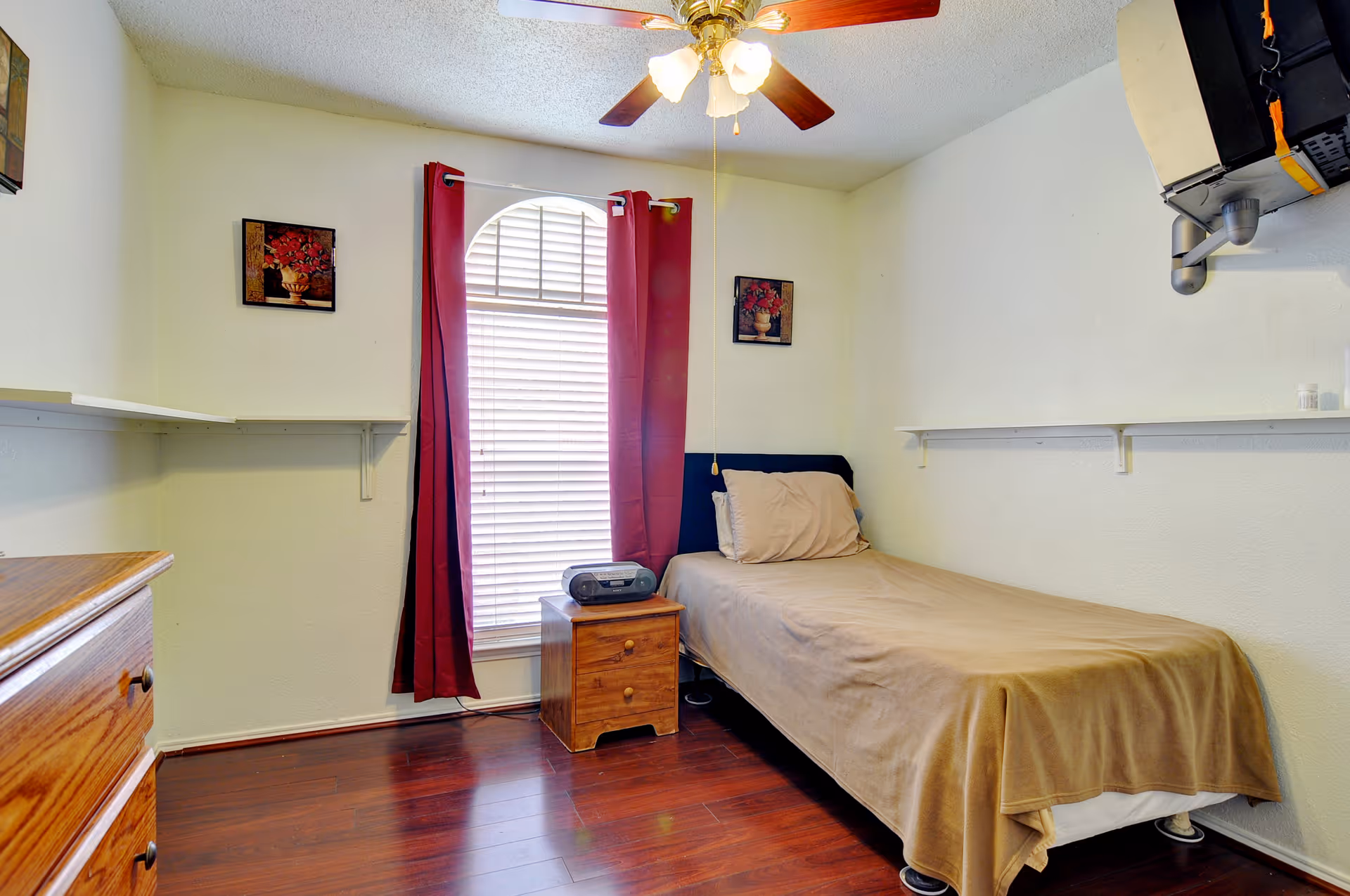 Small single-occupancy bedroom with a twin bed, nightstand, window with red curtains, ceiling fan, and wooden dresser.