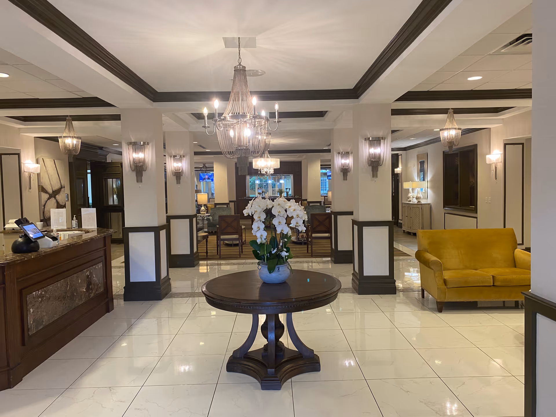 A spacious and elegant interior lobby area with a round wooden table in the center holding a white orchid plant. The floor is tiled with large white tiles, and the ceiling features multiple chandeliers and wall sconces providing warm lighting. To the right, there is a mustard yellow sofa, and to the left, a wooden reception desk with a tablet and hand sanitizer. The background shows more seating areas with chairs and tables.