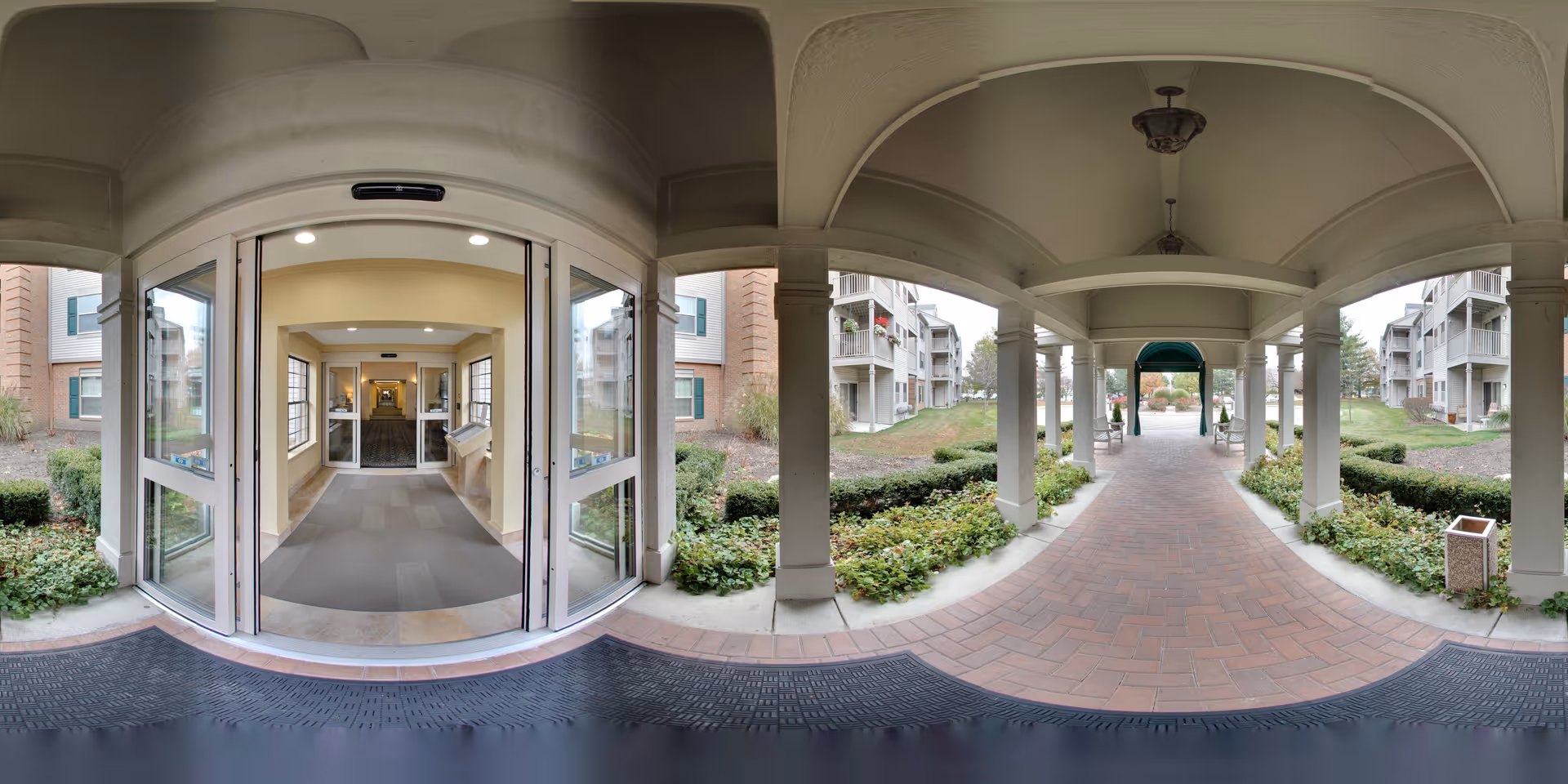 Covered arched entrance with glass automatic doors opening onto a brick walkway and landscaped courtyard flanked by apartment buildings.