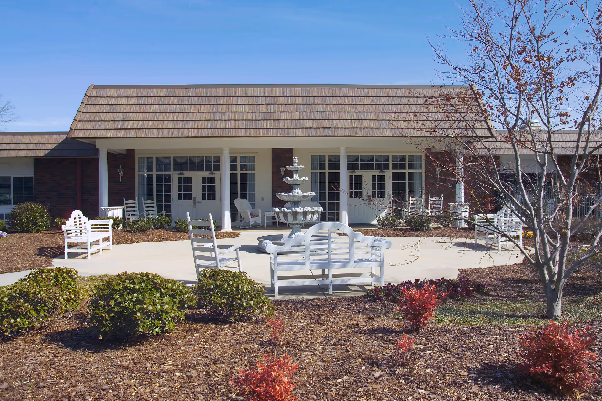 Outdoor patio area of a senior living facility with white wooden benches and rocking chairs arranged around a multi-tiered water fountain. The patio is surrounded by landscaped bushes and small trees, with a building featuring large windows and double doors in the background under a clear blue sky.