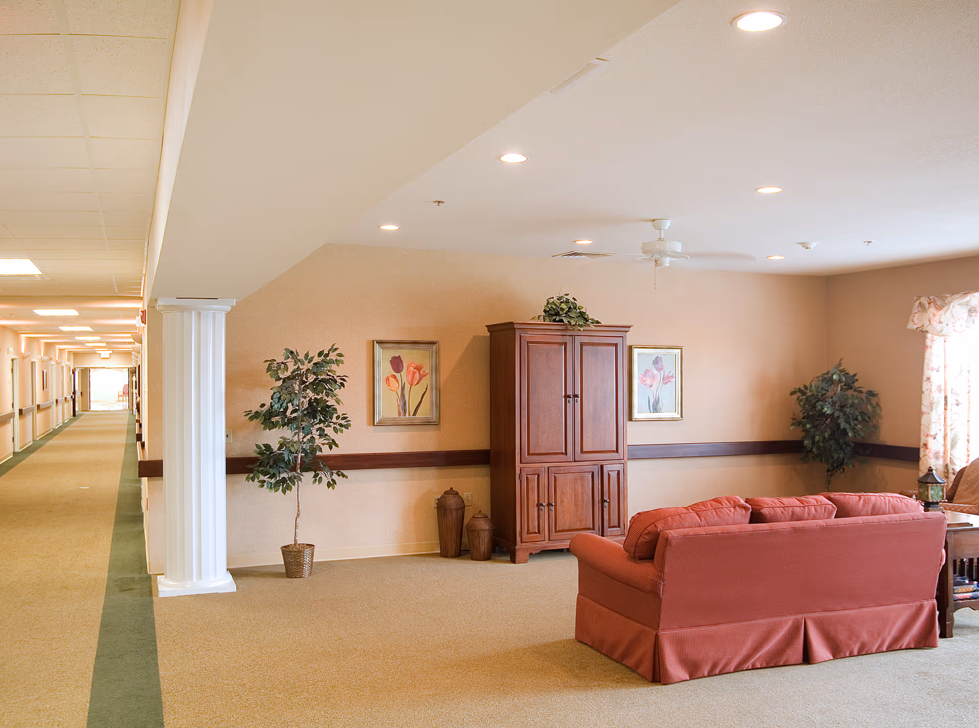 A cozy living room area in a senior living facility with a red sofa, wooden cabinet, two framed floral paintings on the wall, two potted plants, and a window with floral curtains. The room is adjacent to a long hallway with handrails and ceiling lights.