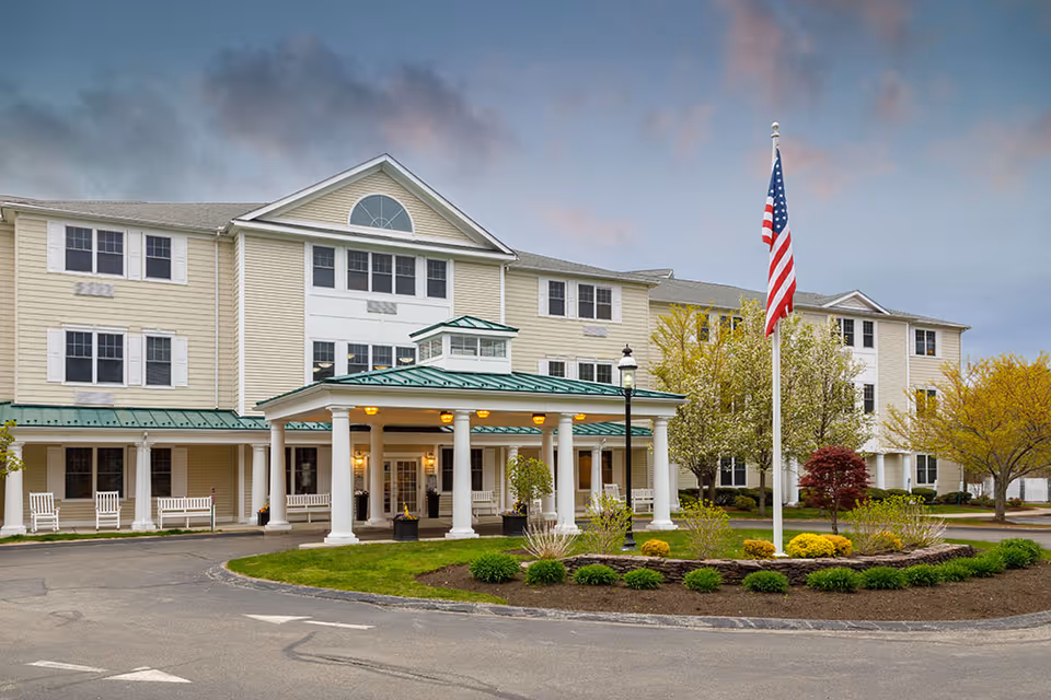 Exterior view of a senior living facility with a three-story beige building featuring white columns and a green roof over the entrance. There are white rocking chairs on the porch, a circular driveway, landscaped garden beds with shrubs and small trees, and an American flag on a flagpole in front of the building under a cloudy sky.