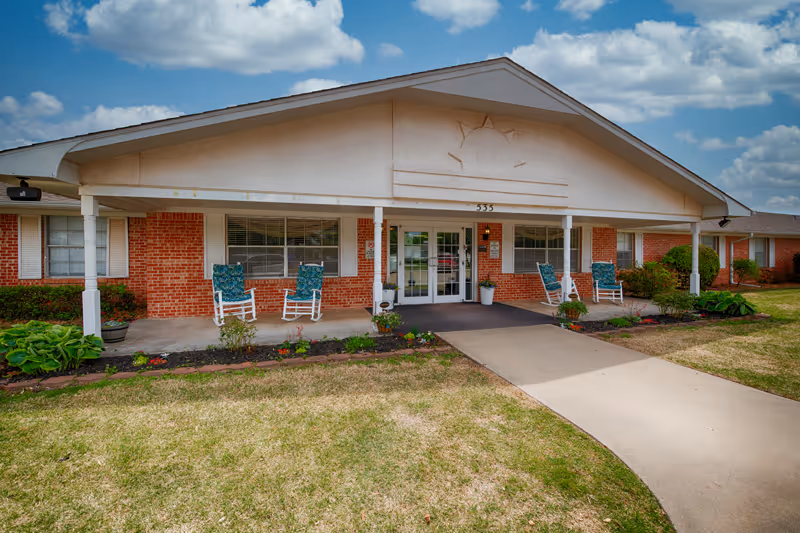 Front exterior view of a single-story brick building with a covered entrance supported by white columns. There are four rocking chairs with blue cushions on the porch, flower beds with plants along the walkway, and a clear sky with scattered clouds above.