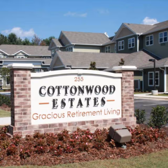 Entrance sign reading "Cottonwood Estates Gracious Retirement Living" with landscaping and the community building in the background.