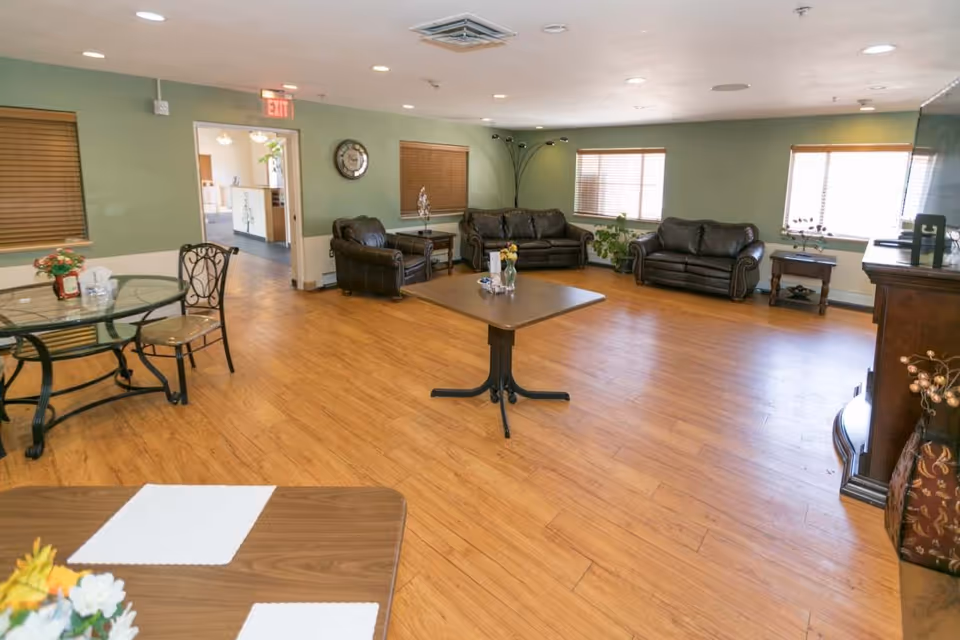 A spacious common living area with wooden flooring, green walls, and multiple windows with blinds. The room features three dark leather sofas arranged around a central wooden coffee table, a glass-top dining table with chairs, and a wooden side table with decorative items. There is an exit sign above a doorway leading to another room.