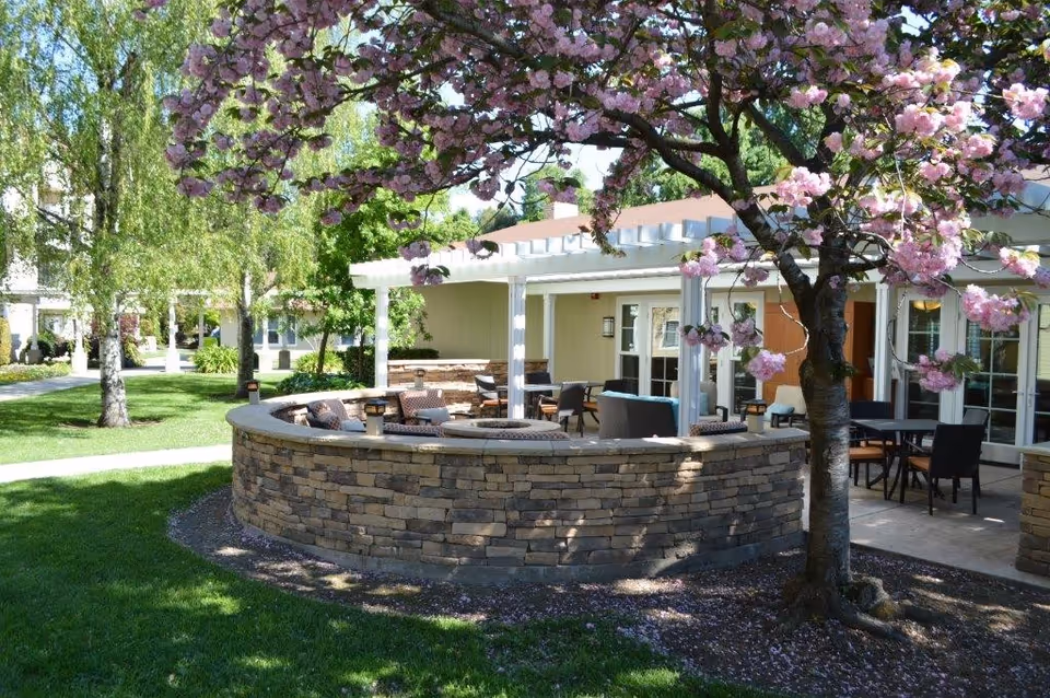 Outdoor seating area with cushioned chairs and tables surrounded by a curved stone wall under a pergola. A tree with pink blossoms is in the foreground, and green grass and other trees are visible in the background.