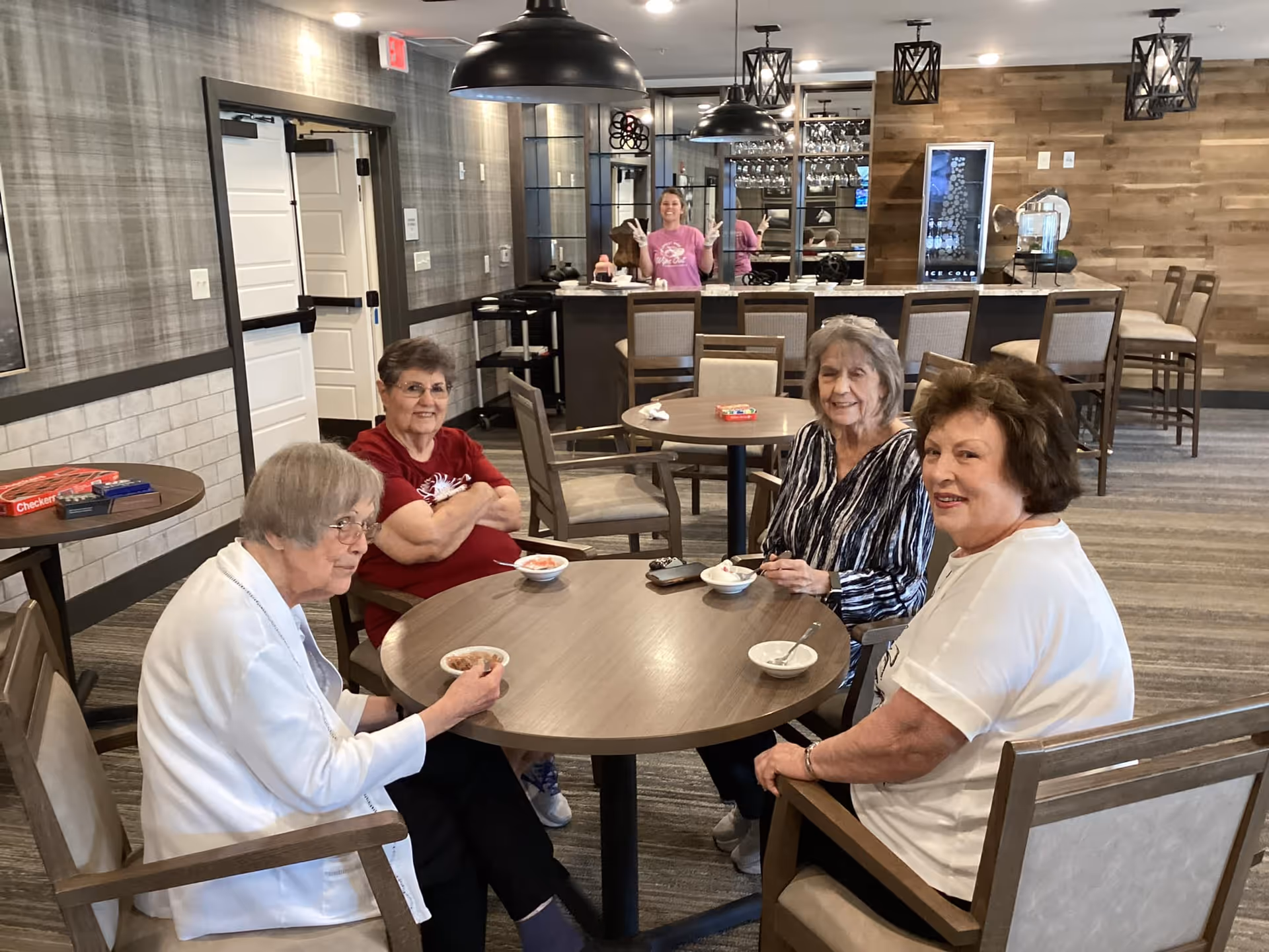 Four elderly women sitting around a round table in a communal dining or lounge area, eating from small bowls. In the background, a woman in a pink shirt is standing behind a counter with bar stools. The room has modern decor with wood paneling, pendant lights, and a carpeted floor.