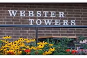 Brick building facade with WEBSTER TOWERS sign and colorful flowers in front.