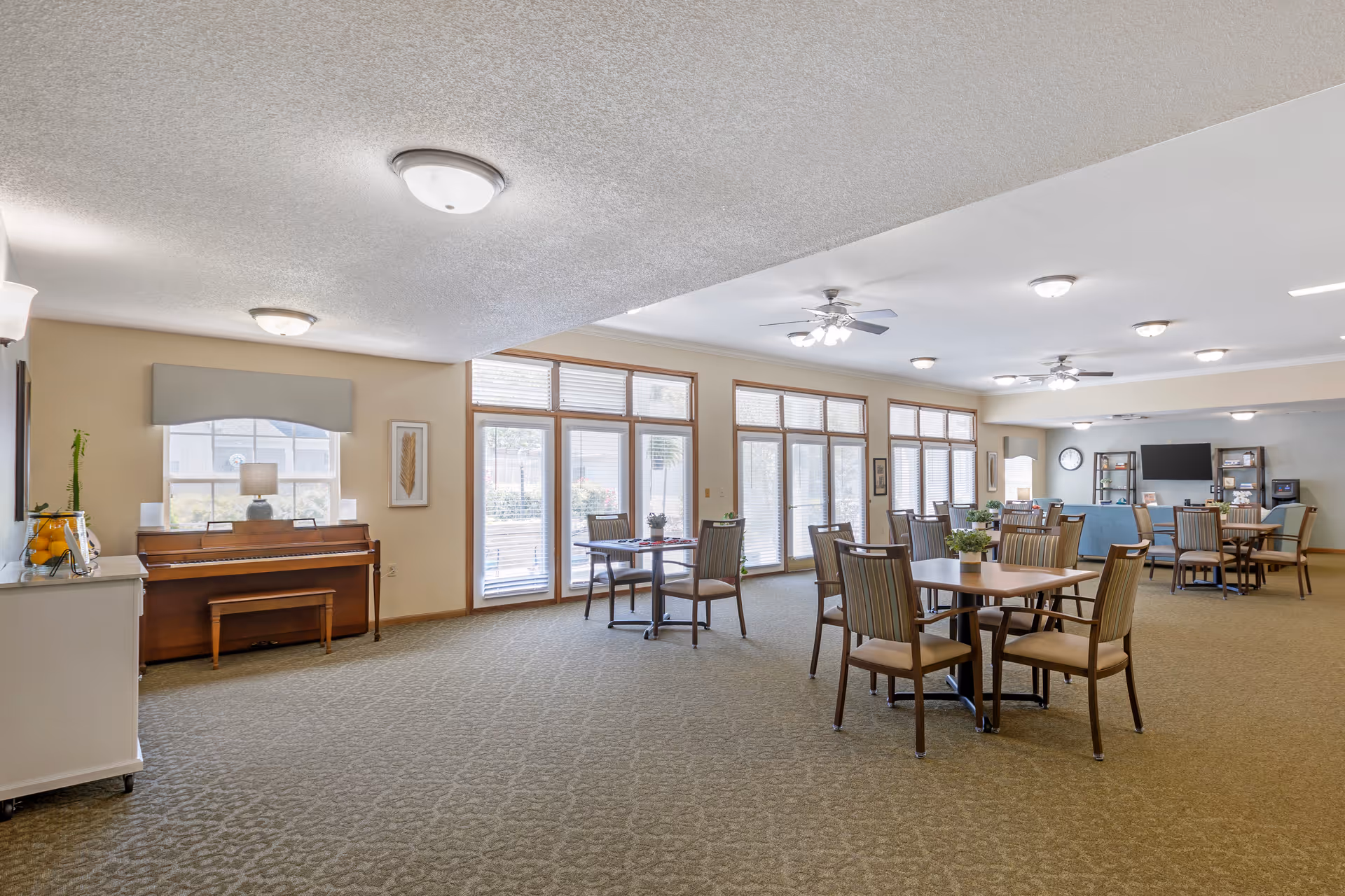 Spacious senior community common room with round dining tables and chairs, a piano, large windows, and a TV lounge area.