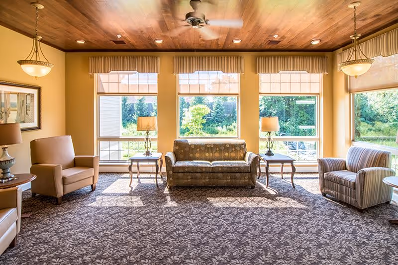 A bright and spacious living room area with large windows letting in natural light. The room features a patterned carpet, a wooden ceiling with a ceiling fan, and three upholstered seating options including a sofa and two armchairs. There are side tables with lamps placed near the windows, and the view outside shows greenery and trees.