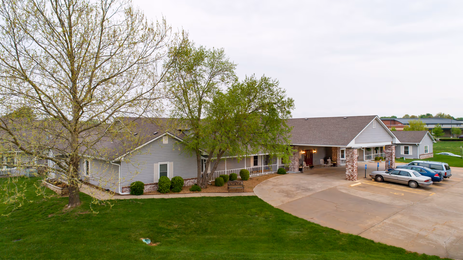 Exterior view of Homestead Assisted Living of Derby, showing a single-story building with a covered entrance supported by brick columns, surrounded by green grass and trees, with a few parked cars in the parking lot.