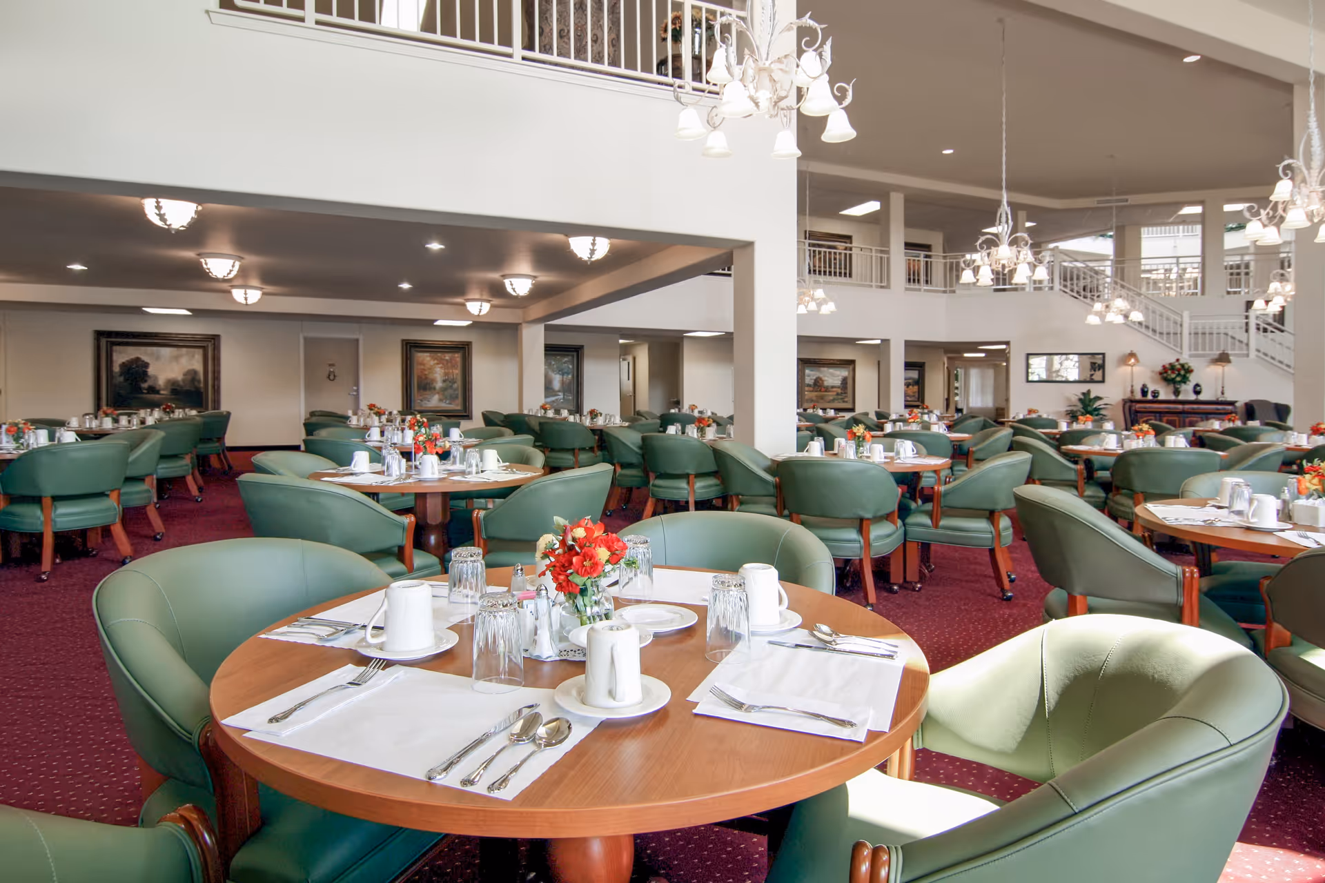 A spacious dining room with multiple round wooden tables set with white placemats, silverware, cups, and glasses. Each table has a small floral centerpiece. The room features green upholstered chairs, red carpet flooring, white walls, and multiple chandeliers hanging from the ceiling. There is a second-floor balcony overlooking the dining area.