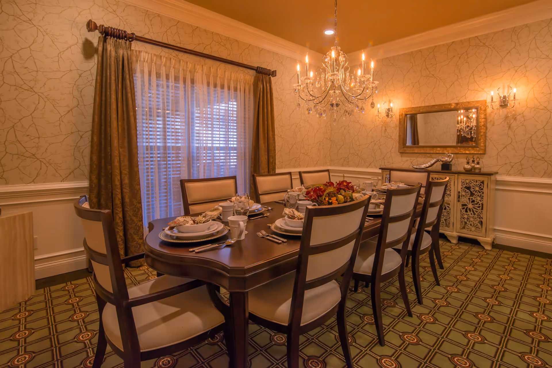A formal dining room with a long dark wood table set for a meal, upholstered chairs, a crystal chandelier, patterned carpet, and a sideboard under a mirror.