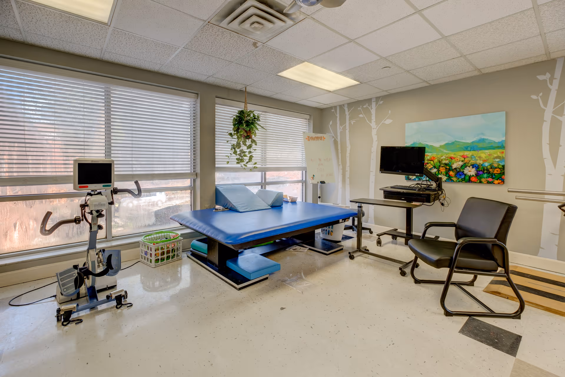 A therapy room with a blue padded treatment table, a stationary exercise bike with a monitor, a black chair, a computer on a rolling desk, and large windows with blinds. The walls are decorated with white tree decals and a colorful landscape painting.