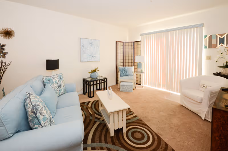 A bright living room with a light blue sofa adorned with floral pillows, a white coffee table on a patterned rug, a white armchair with a pillow, a small side table with a lamp, and a folding screen near vertical blinds covering a large window or sliding door.