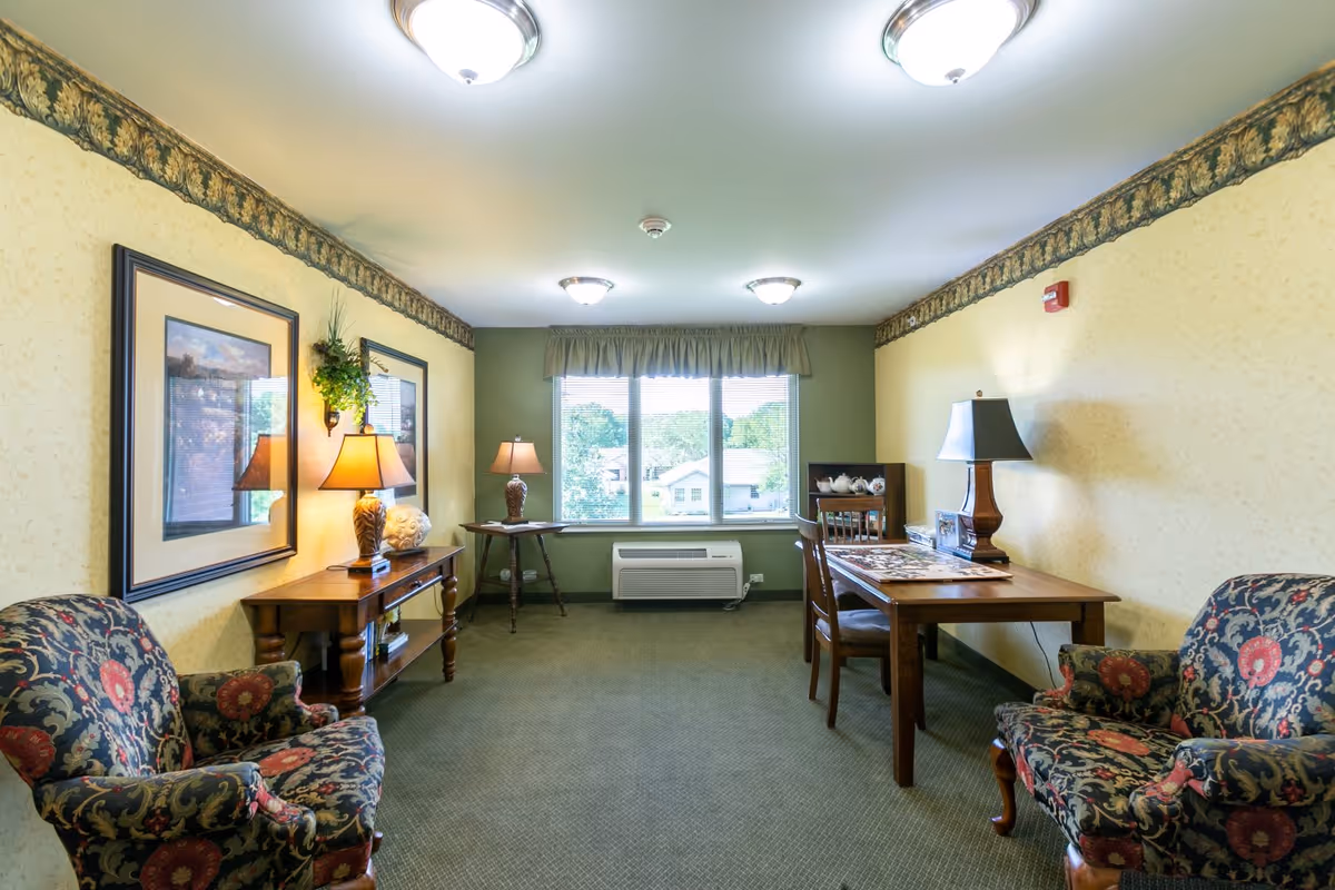 A cozy sitting room with two patterned armchairs facing each other, a wooden table with a lamp and a puzzle on it, a wooden console table with a lamp and decorative items, two framed pictures on the wall, and a large window with a view of houses and trees outside.