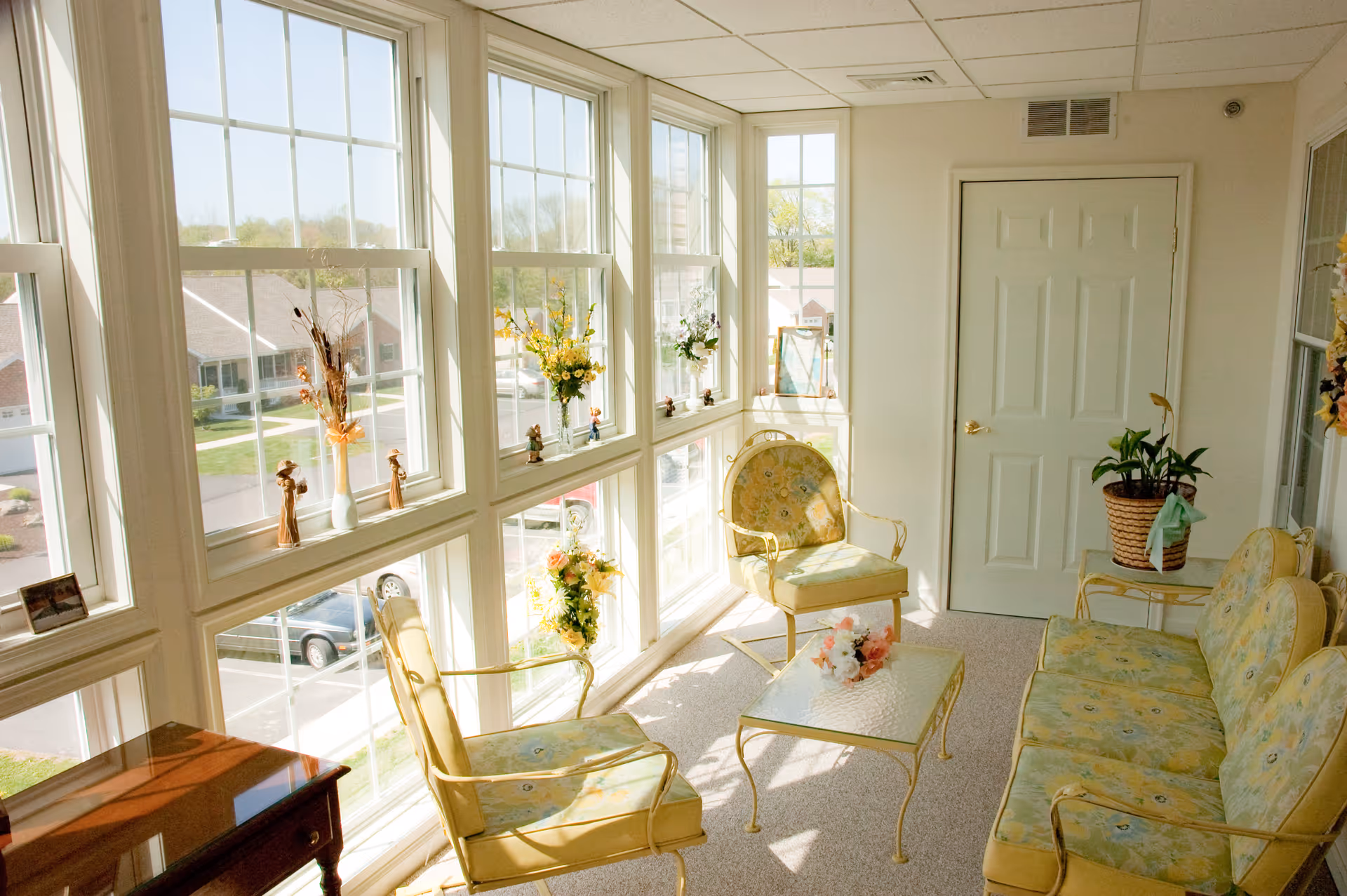 Sunlit sunroom-style sitting area with multiple large windows, floral-upholstered chairs and a small glass coffee table.